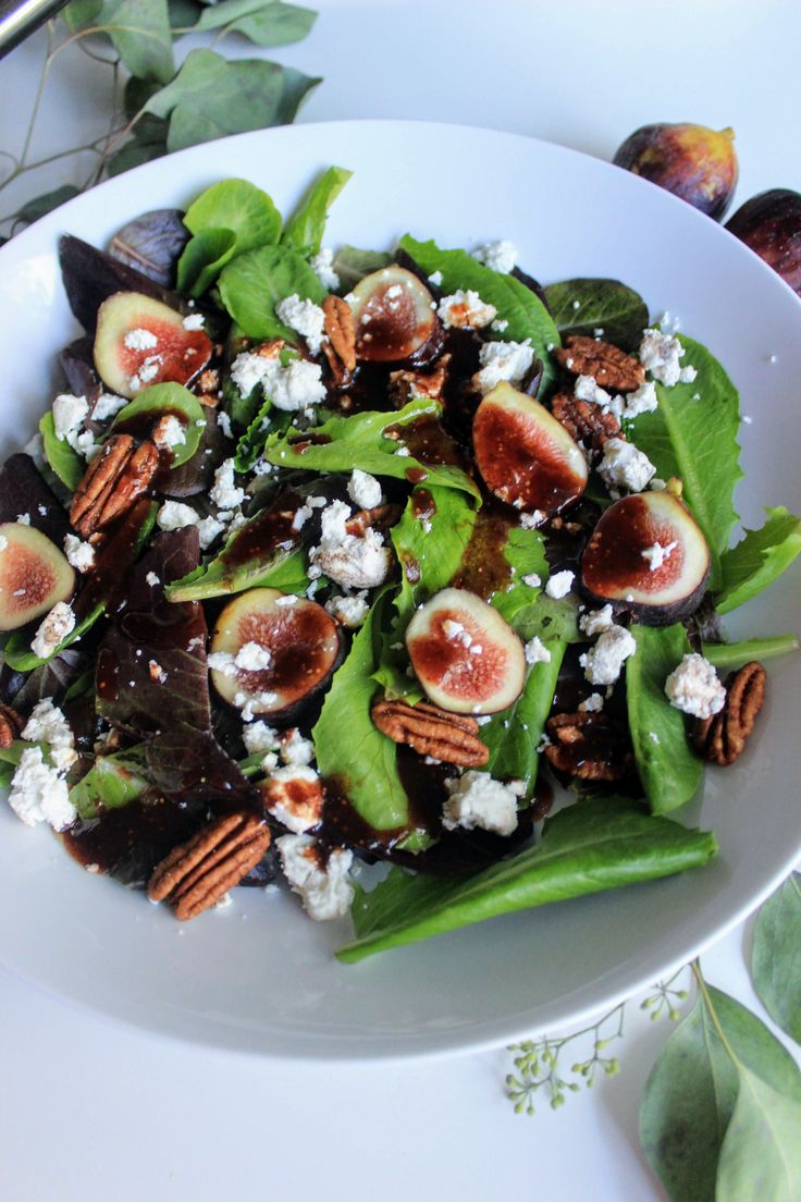 a salad with figs, cheese and nuts on a white plate next to green leaves