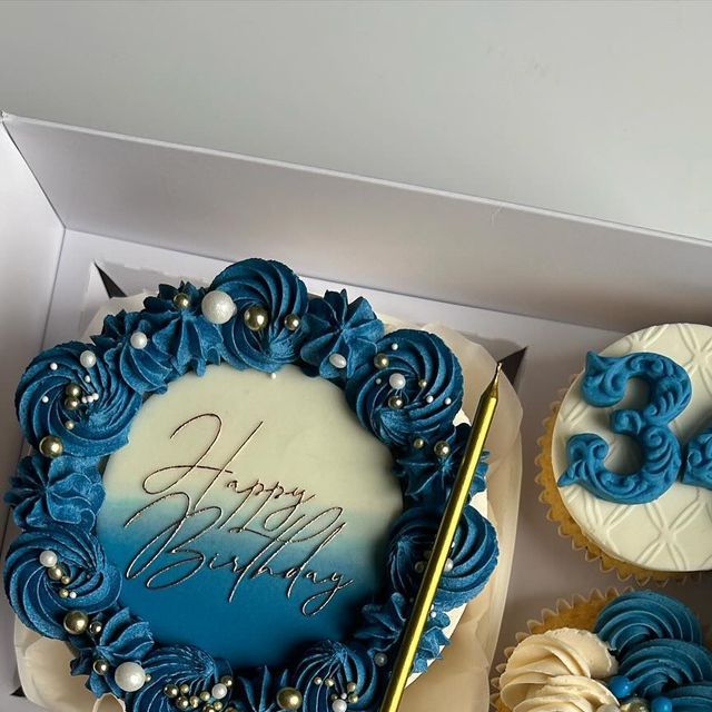 a blue and white birthday cake in a box with cupcakes next to it