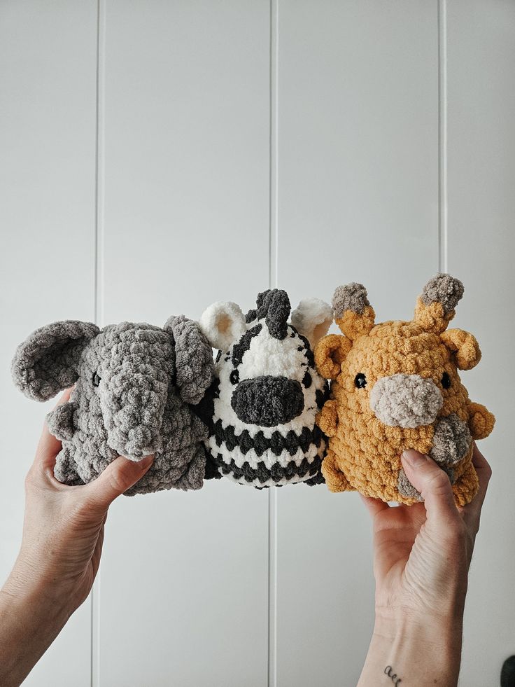 three crocheted stuffed animals are being held by two hands in front of a white wall