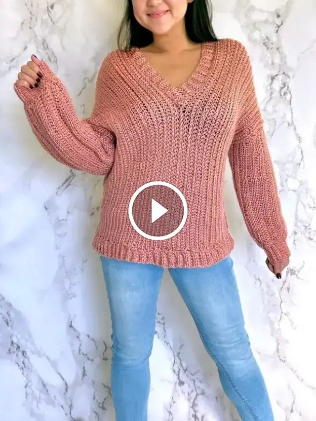 a woman standing in front of a marble wall wearing a pink sweater and blue jeans