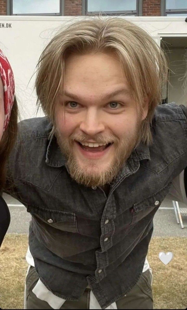 a man with long hair and beard standing in front of a building smiling at the camera