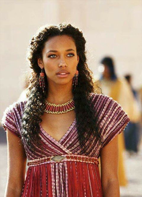 a woman with long hair wearing a red dress and gold jewelry on her neck is looking at the camera