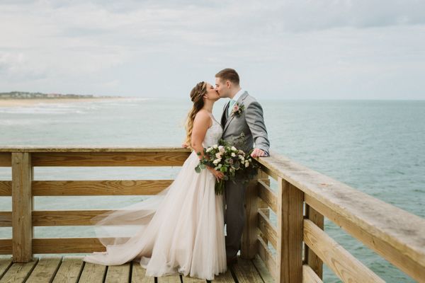 a bride and groom kissing on a pier overlooking the ocean