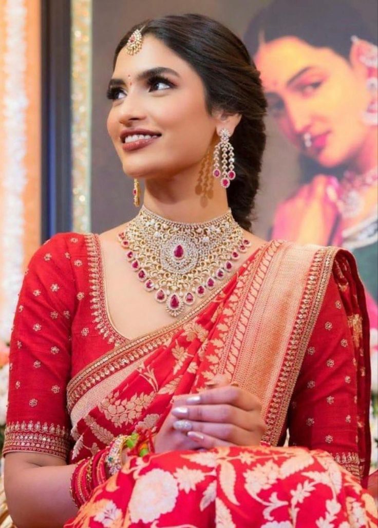 a woman in a red and gold sari with jewelry on her neck, smiling at the camera