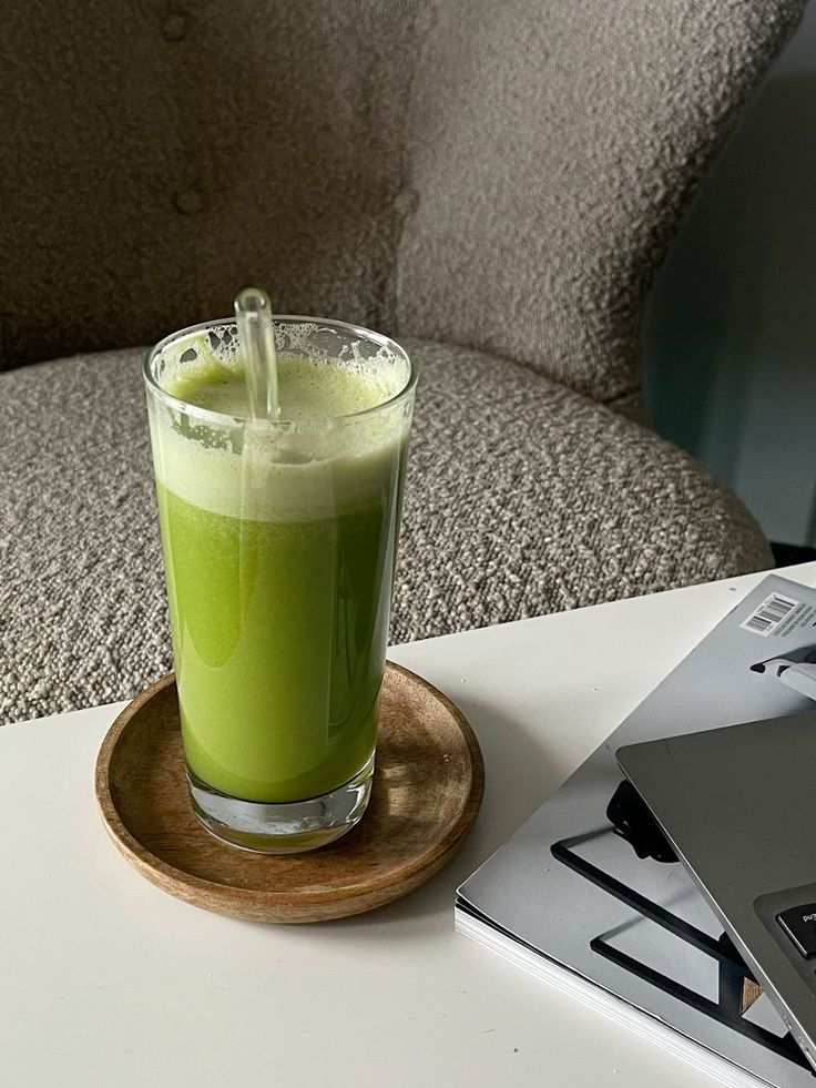 a glass filled with green liquid sitting on top of a white table next to a laptop