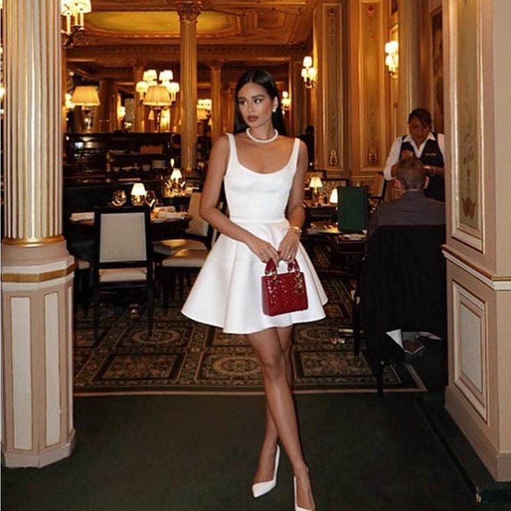 a woman in a short white dress is holding a red purse and posing for the camera