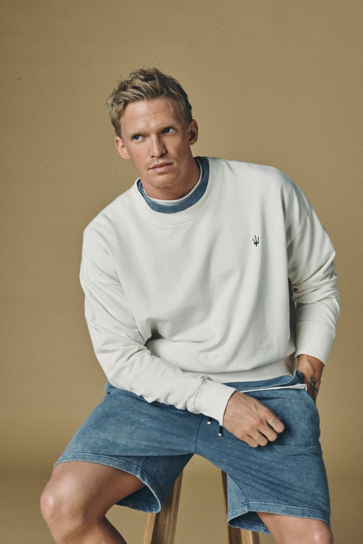 a man sitting on top of a wooden stool wearing a white sweater and blue shorts