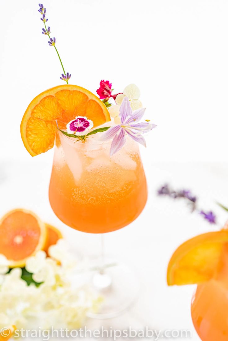 an orange cocktail garnished with fresh flowers and lavender sprigs on a white table