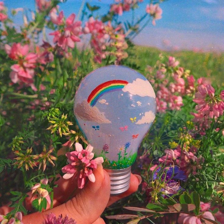 a hand holding a light bulb with a rainbow painted on it in the middle of flowers