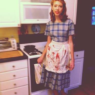 a woman standing in front of an oven with blood all over her dress and apron