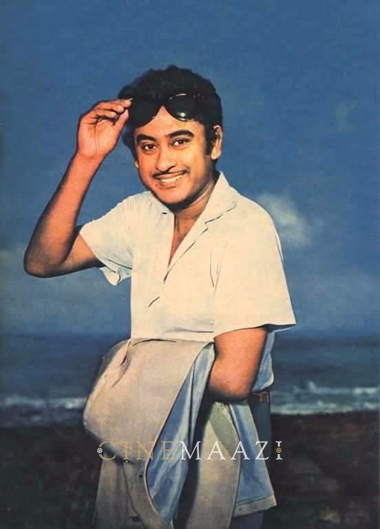 a man in white shirt standing on beach next to the ocean with sunglasses on his head