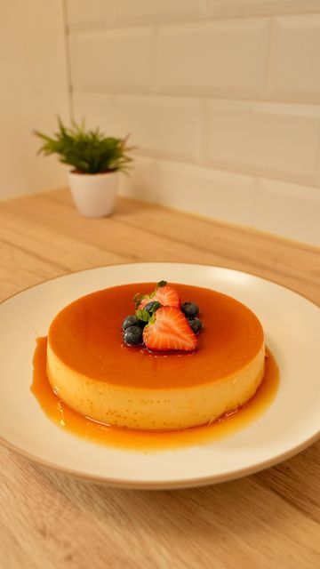 a small cheesecake on a white plate topped with berries and sauce next to a potted plant