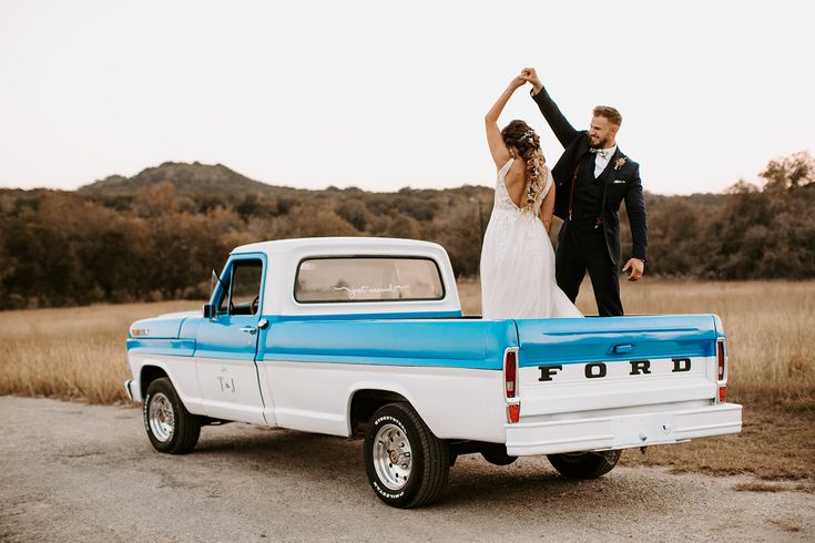 a bride and groom standing in the back of a pickup truck with their arms raised