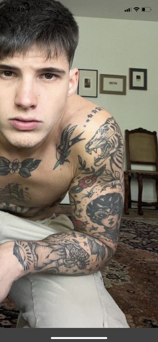 a man with tattoos on his arms and chest sitting in front of a rug looking at the camera