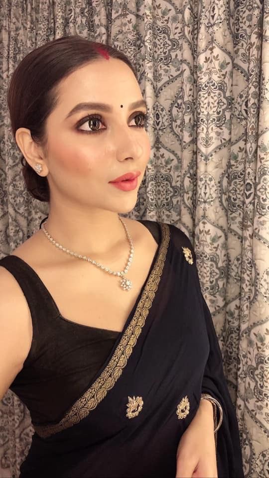 a woman wearing a black sari with gold accents and pearls on her neck, standing in front of a curtain