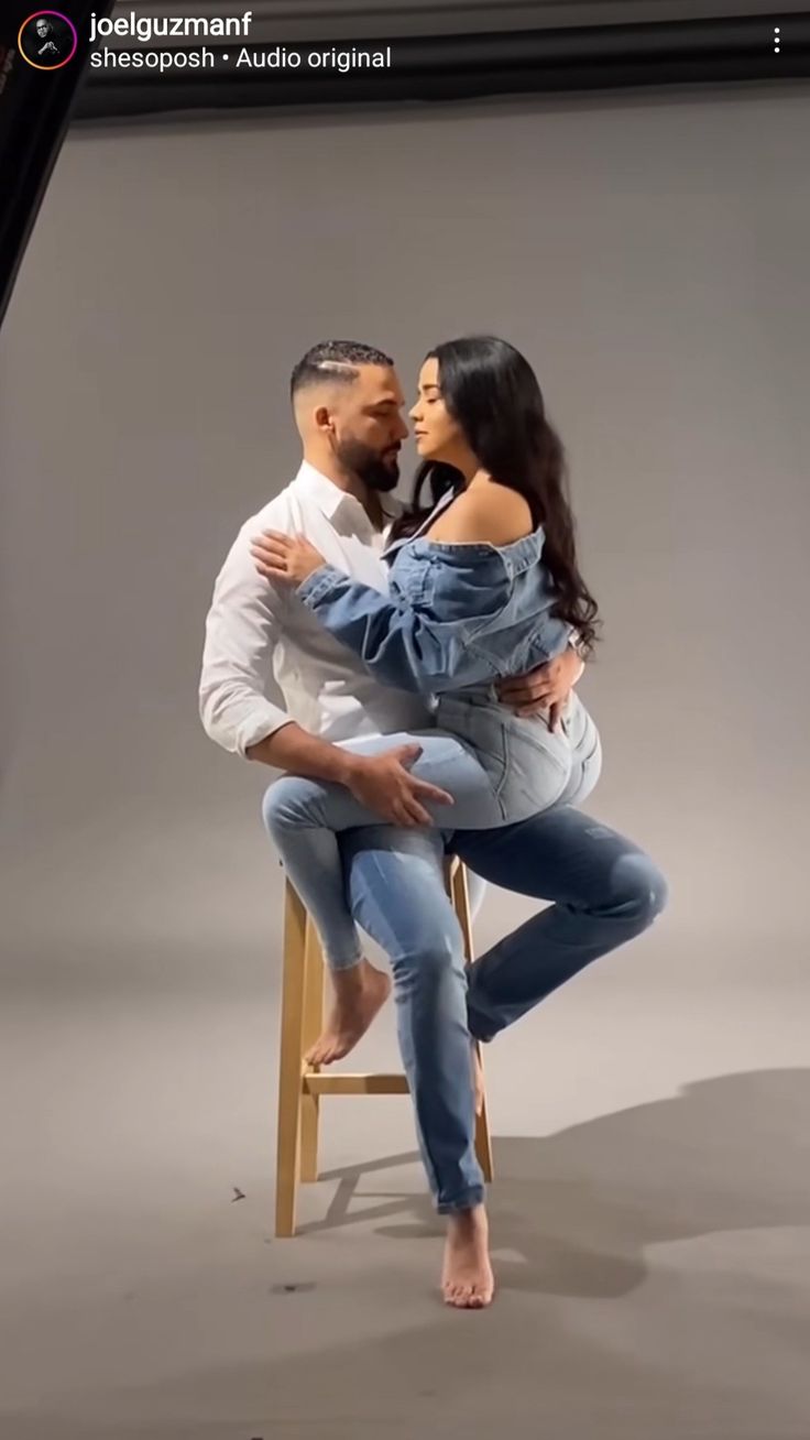 a man and woman sitting on a chair in front of a gray background, posing for the camera