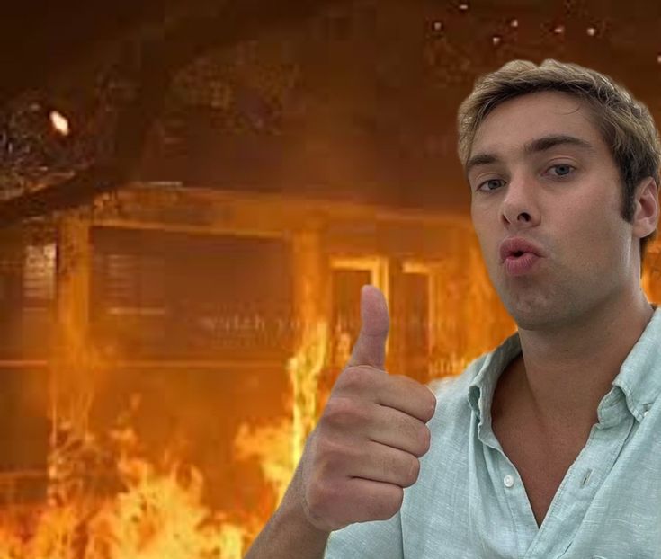 a man giving the thumbs up in front of a house on fire with flames behind him