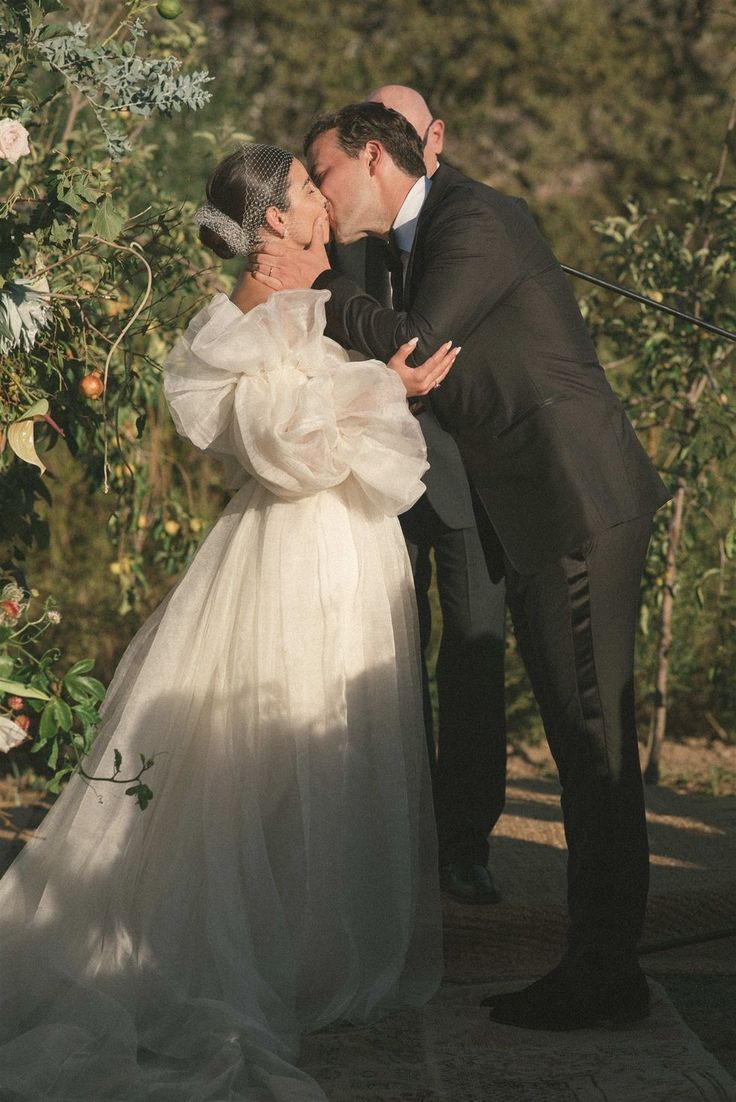 a bride and groom kissing in front of an arch with flowers on it's side