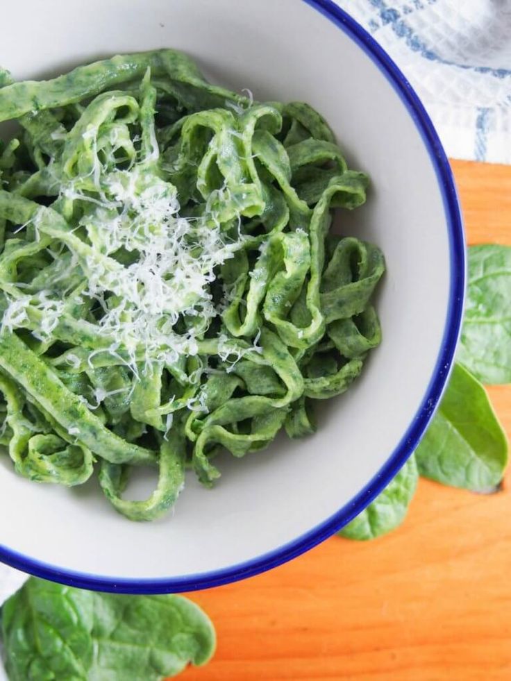 a white bowl filled with green pasta and spinach on top of a wooden table