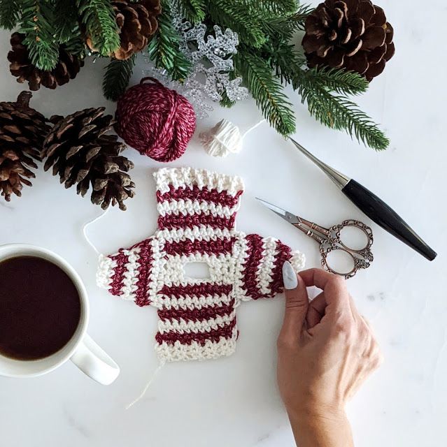 someone is crocheting a knitted sweater next to a cup of coffee and pine cones