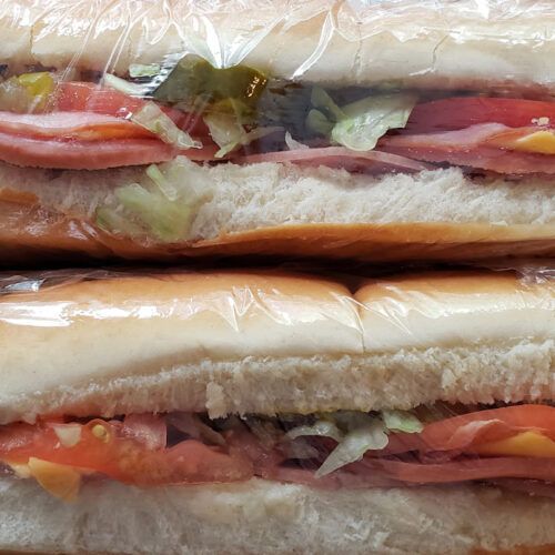 two sub sandwiches wrapped in plastic sitting on top of each other with meat and veggies