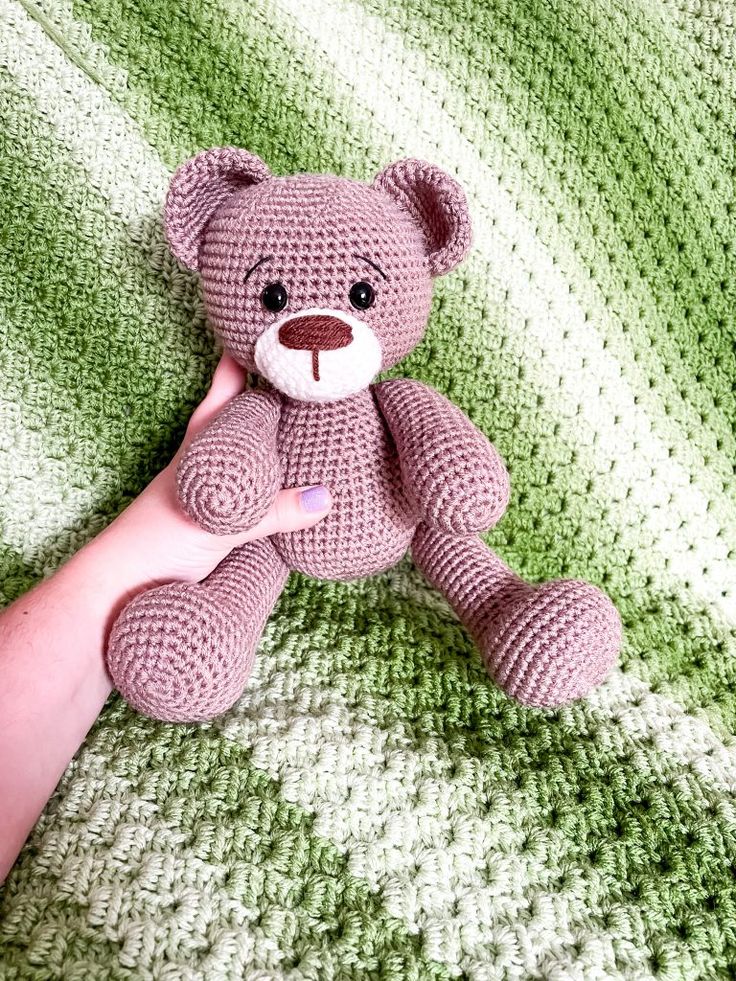 a hand holding a pink crocheted teddy bear on top of a green blanket