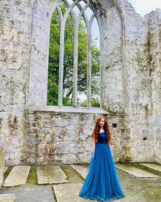 a woman standing in front of an old stone building wearing a blue dress and looking at the camera