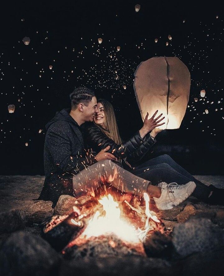 a man and woman sitting next to a campfire with lanterns in the sky behind them