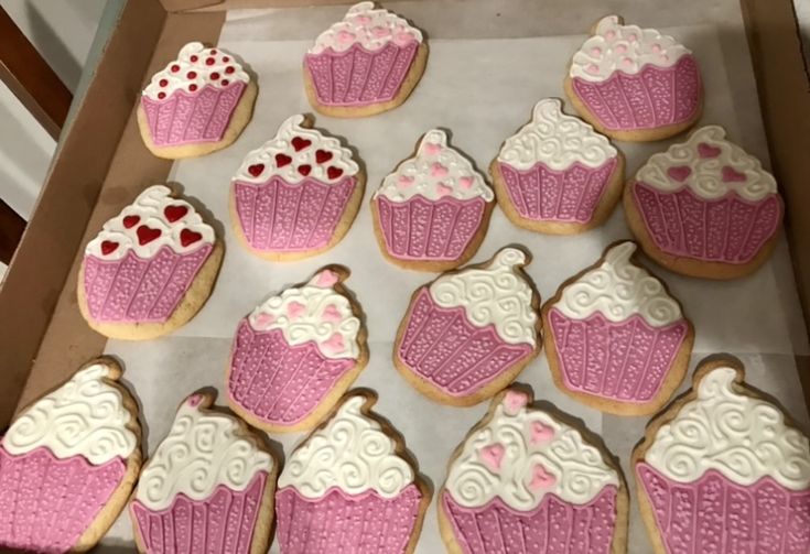 pink and white decorated cupcakes are in a box on the table, ready to be eaten