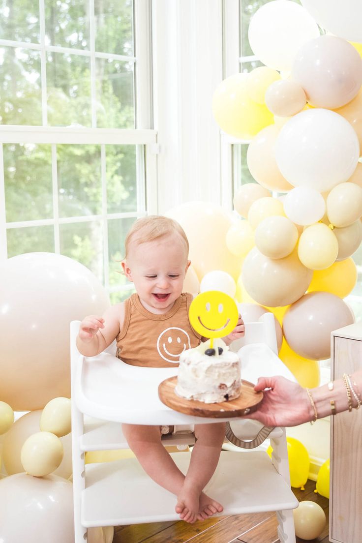 a baby is sitting in a high chair with a cake and balloons behind him that are yellow and white