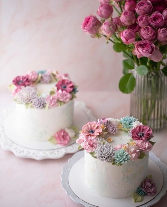 two cakes decorated with flowers on plates next to a vase filled with pink and purple roses