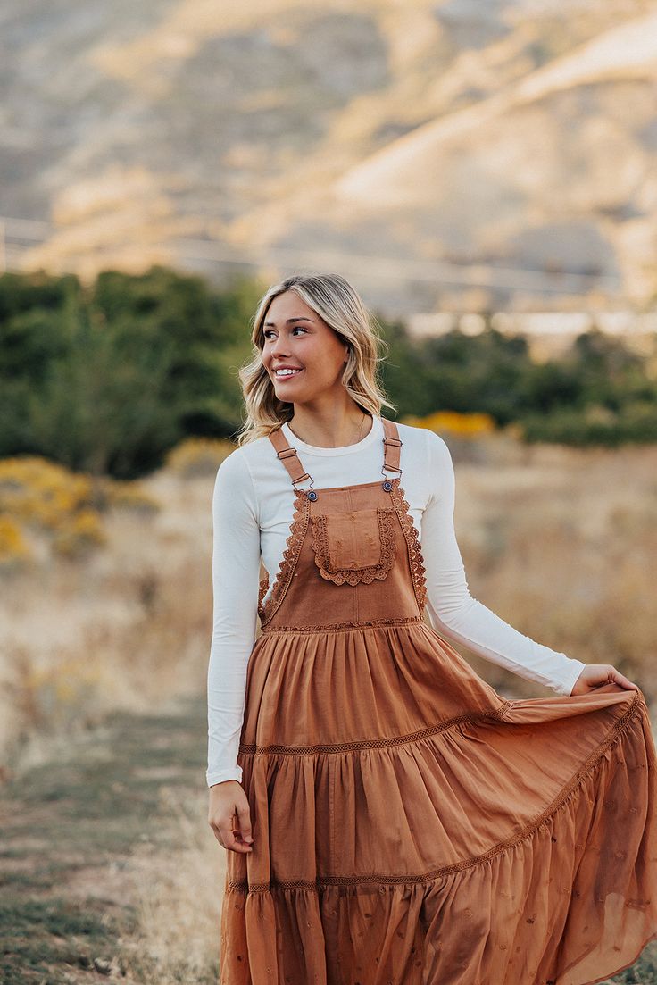 Step out in style with THE EMILIE LACE SKIRTALL IN RUST. This overall dress is sure to bring you plenty of compliments with its rust hue, chic lace detailing, and chiffon contrast panel. Don't forget to twirl in this one-of-a-kind midi overall dress! Paired with THE LAYERING CAP SLEEVE TEE IN WHITE BY MIKAROSE and THE FLORANCE FLORAL MESH LAYERING LONG SLEEVE IN BIEGE Embroidered lace details Midi length Chiffon contrast panel Tiered Self: 100% cotton, Contrast: 100% polyester, Lining: 100% rayon Runs true to size. Small (2-4), Medium (4-8), Large (8-12). Length (from shoulder to hem): Small: 51", Medium: 51.5", Large: 52". Model is 5'8" wearing a size small. To view our return policy, click here! Check out our try on video below! Overall Dress Outfit Fall, Fall Senior Pics Outfits, Heartland Outfits, Long Sleeve Shirt Under Dress, Western Dress Outfits, Layered Dress Outfit, Overall Dress Outfit, Mesh Layering, Layering Long Sleeve