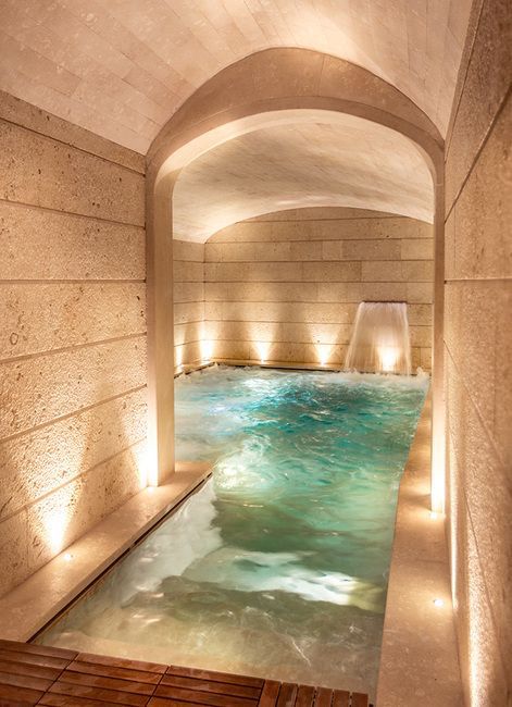 an indoor swimming pool surrounded by stone walls and wooden flooring, with lights on the side