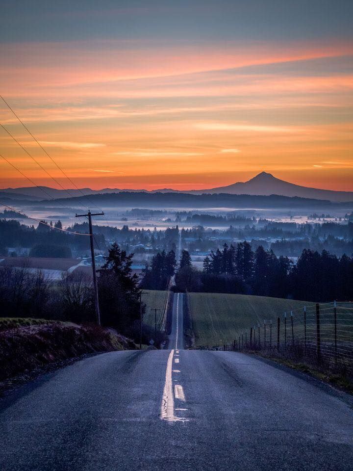 an empty road with mountains in the distance at sunset or sunrise, as seen from across the street