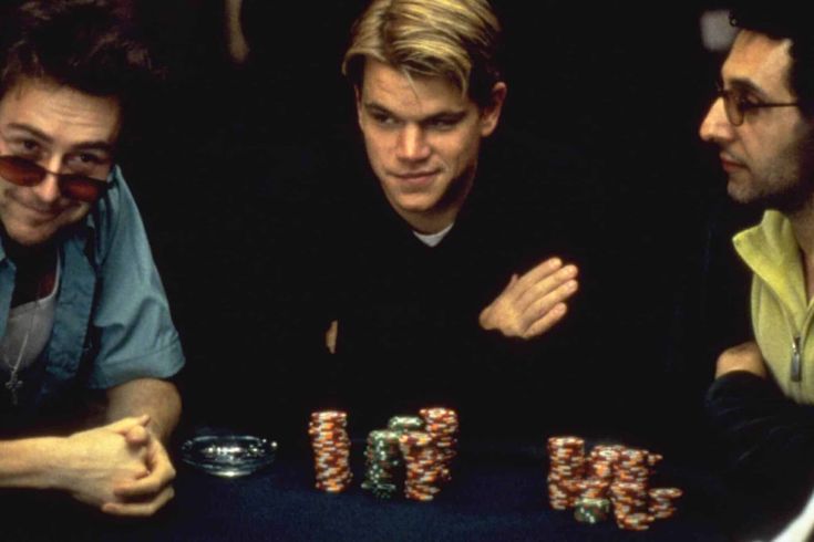 three men sitting at a table with poker chips in front of them and one man holding his hand up