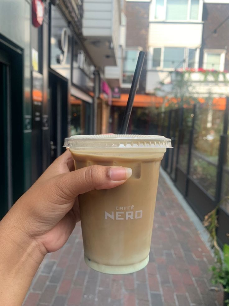 a hand holding up a coffee cup with a straw in it on a sidewalk next to buildings