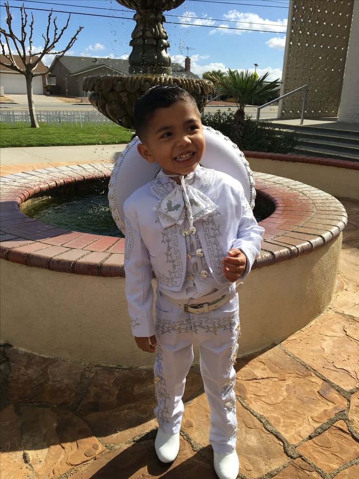 a little boy dressed in white standing next to a fountain with his hands on his hips