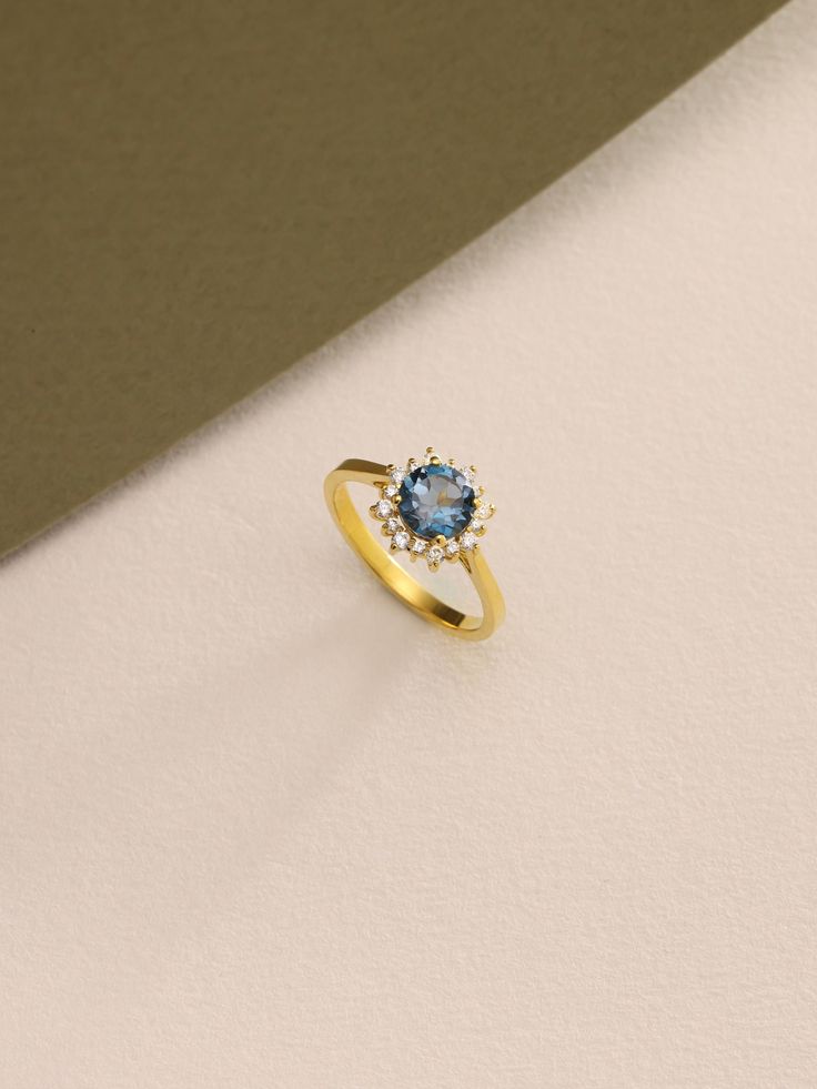 a gold ring with a blue topazte and white diamonds on the side, against a beige background