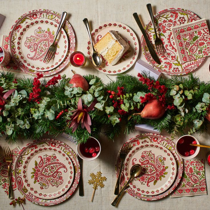 the table is set for christmas dinner with red and white plates, silverware, holly wreaths, candles, and fruit