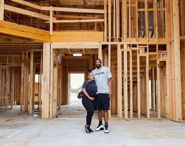 a man and woman standing in an unfinished building with wooden framing on the walls, looking at the camera