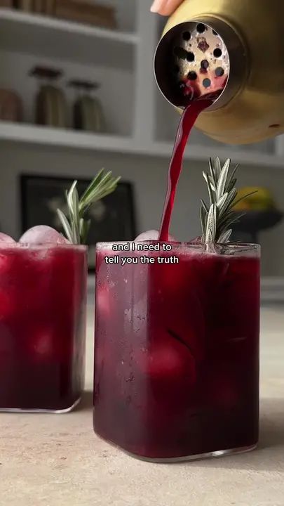 someone pours red liquid into two glasses filled with ice and rosemary sprigs