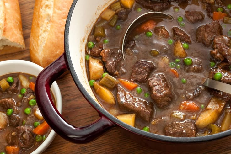 a bowl of beef stew with peas and carrots next to a loaf of bread