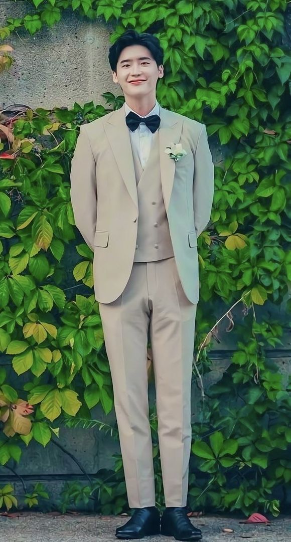 a young man in a suit and bow tie standing next to a wall covered with green plants