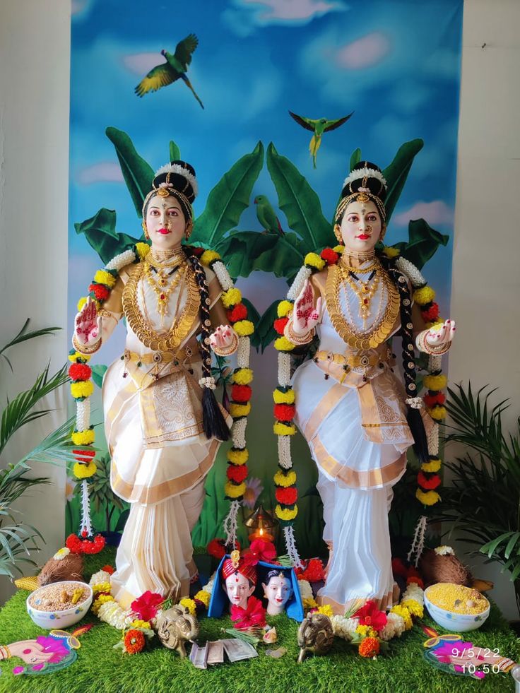 two statues of deities in front of a wall with flowers and birds on it, surrounded by greenery