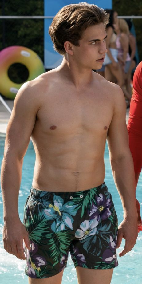 a shirtless young man standing in front of a swimming pool with people around him