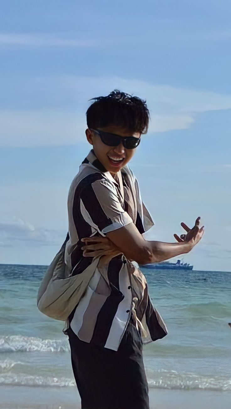 a young man standing on top of a sandy beach next to the ocean with his arms crossed