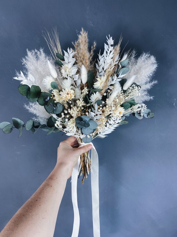 a hand holding a bouquet of flowers with white and green foliage on it, against a blue background