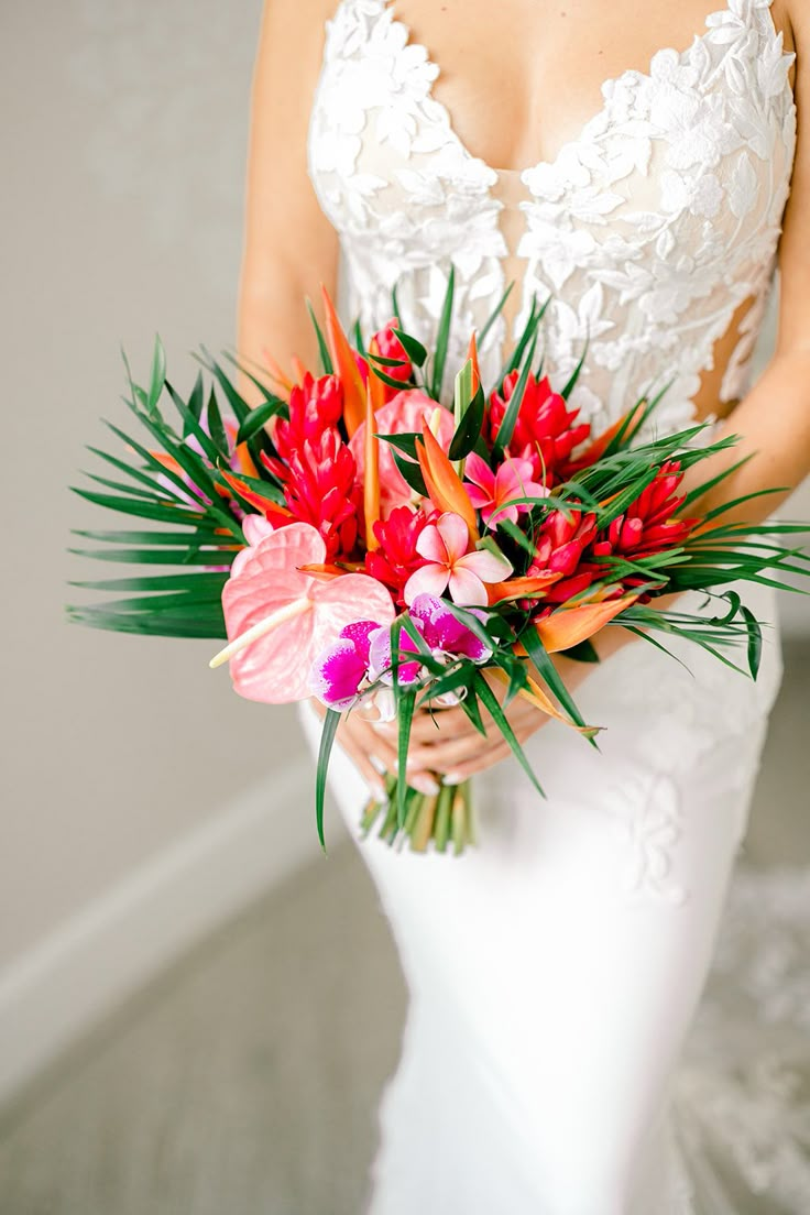 a woman in a wedding dress holding a bouquet of red and pink flowers with palm leaves