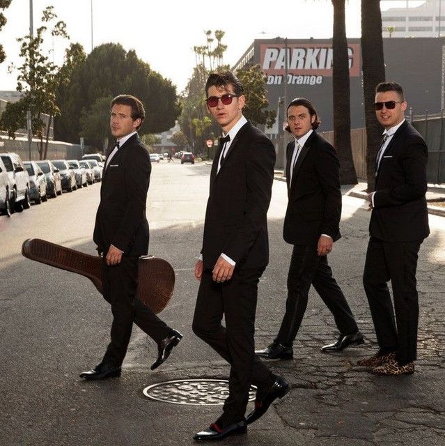 three men in suits and sunglasses walking down the street with a large piece of luggage