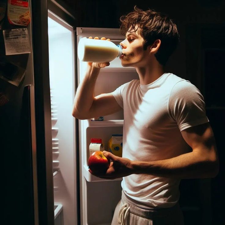 a young man drinking from a bottle while standing in front of an open refrigerator door